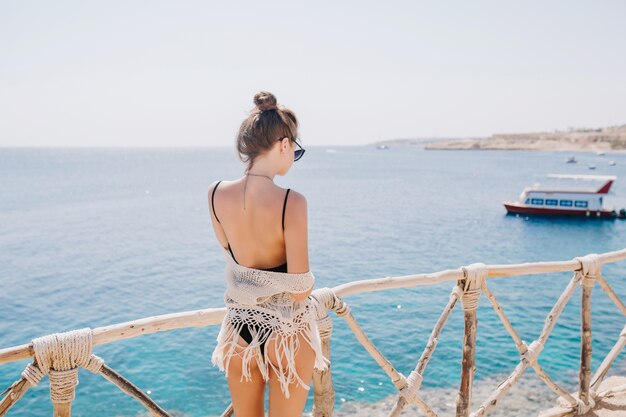 Chica delgada y elegante con peinado de moda disfrutando de vistas al mar y mirando el lanzamiento. Retrato de espalda de increíble joven en traje de baño negro de pie en el océano