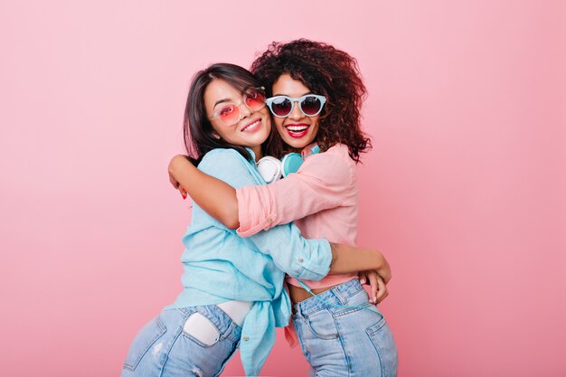 Chica delgada emocionada con peinado africano abrazando a una amiga asiática en gafas de sol coloridas de moda. Señora bastante europea en jeans abraza a mujer joven negra con camisa rosa.