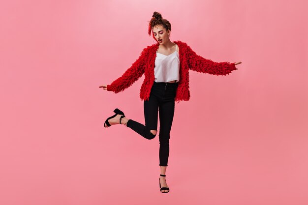 Chica delgada en jeans oscuros y abrigo rojo bailando en rosa