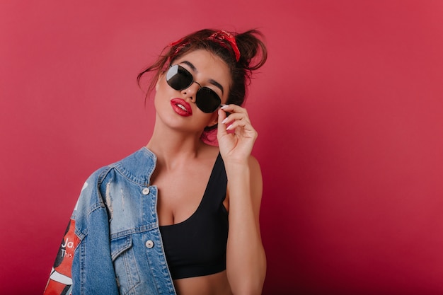 Foto gratuita chica delgada con labios rojos de pie en el espacio oscuro y tocando gafas de sol negras