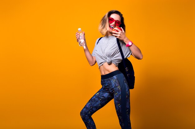 Chica delgada con pelo corto con mochila yendo al gimnasio y sosteniendo una botella de agua. Riendo a joven deportiva con gafas de sol posando sobre fondo brillante en estudio con manzana en mano.