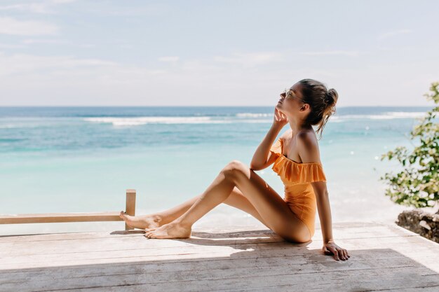 Chica delgada pensativa sentada en el suelo con los ojos cerrados y escuchando las olas del mar. Tiro al aire libre de hermosa mujer europea posando en la costa del mar.