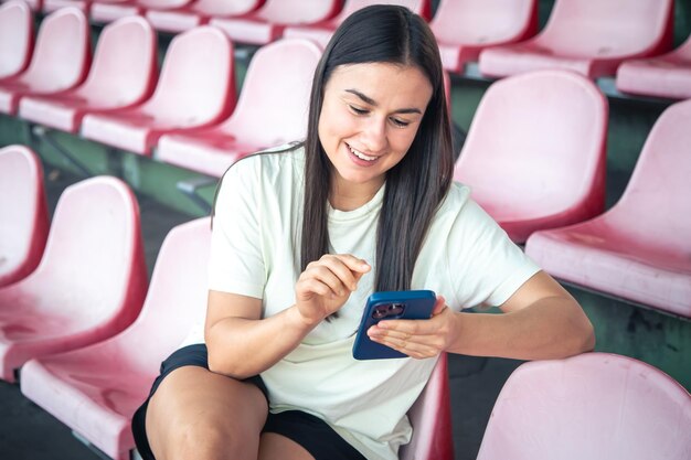 Foto gratuita chica delgada con ropa deportiva sentada en los asientos del estadio y usando un teléfono inteligente