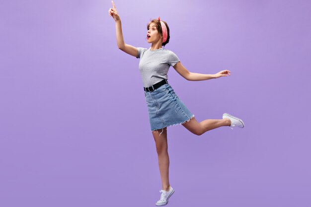 Chica delgada en traje de mezclilla posando.n fondo morado. Señorita sorprendida con pañuelo rosa en zapatillas blancas y camisa de moda saltando.