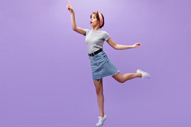 Chica delgada en traje de mezclilla posando.n fondo morado. Señorita sorprendida con pañuelo rosa en zapatillas blancas y camisa de moda saltando.