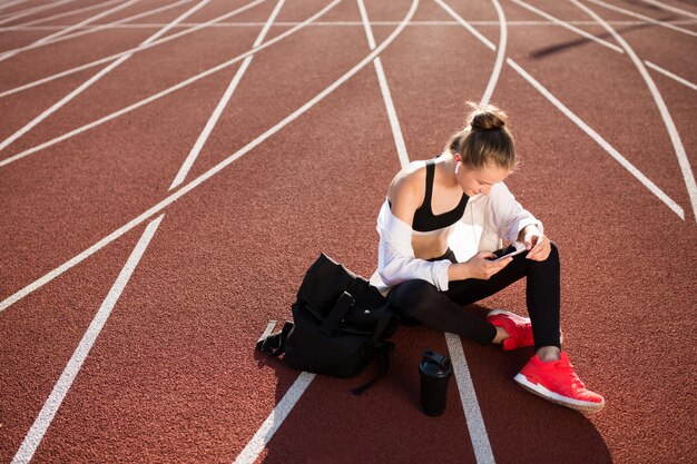 Chica deportiva en auriculares inalámbricos con mochila y botella deportiva cerca felizmente usando el teléfono celular mientras pasa tiempo en la cinta de correr del estadio