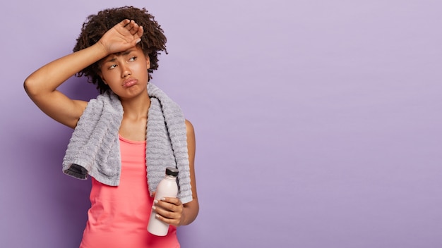 Foto gratuita chica deportiva con cabello afro se limpia la frente, está sudada, vestida con un chaleco informal, sostiene una botella con agua fresca, tiene entrenamiento regular para mantenerse en forma, usa una toalla en los hombros