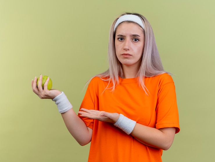 Foto gratuito chica deportiva caucásica joven despistada con tirantes con diadema y muñequeras sostiene y apunta a apple