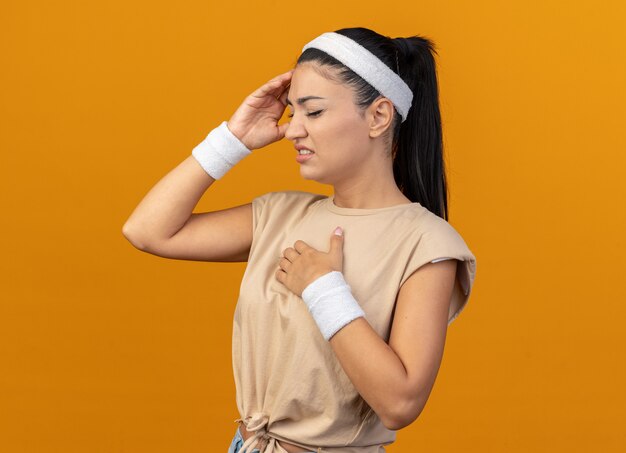 Chica deportiva caucásica joven dolorida con diadema y muñequeras de pie en la vista de perfil manteniendo la mano en el pecho tocando la cabeza con los ojos cerrados aislados en la pared naranja con espacio de copia