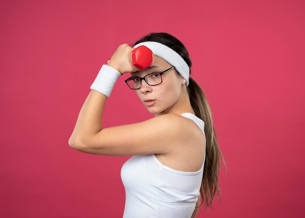 Chica deportiva joven confiada en gafas ópticas con diadema y muñequeras se encuentra de lado sosteniendo mancuernas