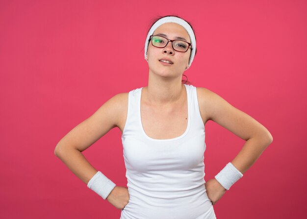 Chica deportiva joven confiada en gafas ópticas con diadema y muñequeras pone puños en la cintura y mira a la cámara