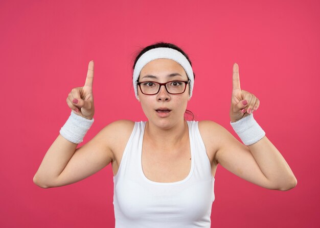 Chica deportiva joven sorprendida en gafas ópticas con diadema y muñequeras apunta hacia arriba con las dos manos