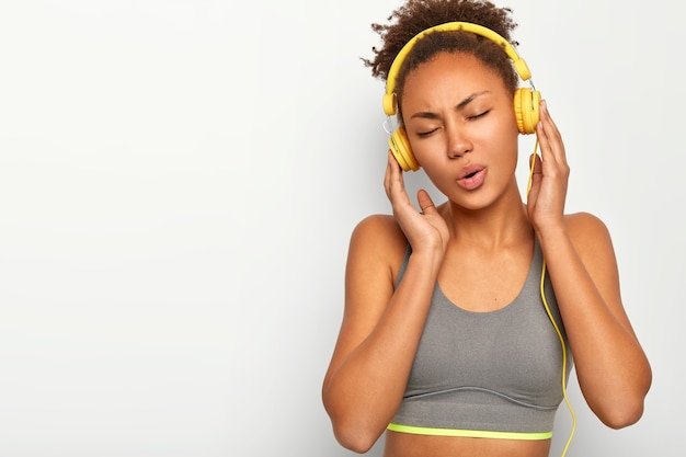 Chica deportiva de pelo rizado relajado, hace ejercicio en el interior, canta canciones, escucha música en auriculares, viste una camiseta gris