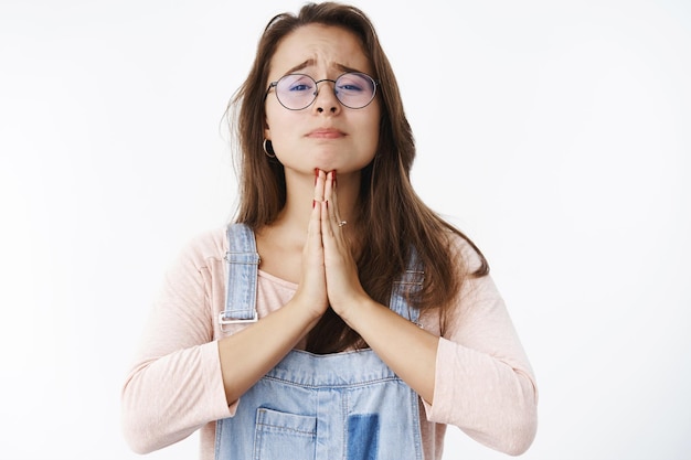 Chica desesperada que tiene problemas para pedir ayuda mientras se toma de la mano en oración con el ceño fruncido y se ve nerviosa, pidiendo un favor, estando en una situación problemática posando sobre una pared gris