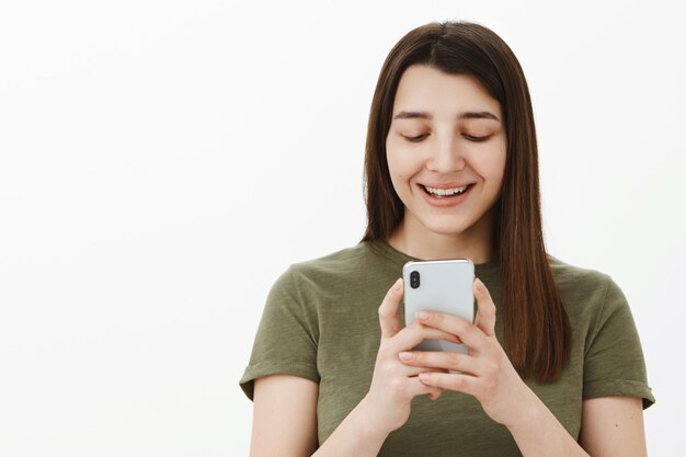 Chica desplazándose memes divertidos a través de un teléfono inteligente riendo y divirtiéndose sosteniendo el teléfono móvil cerca de la cara sonriendo y luciendo divertida y complacida en la pantalla del teléfono celular posando encantada contra la pared blanca