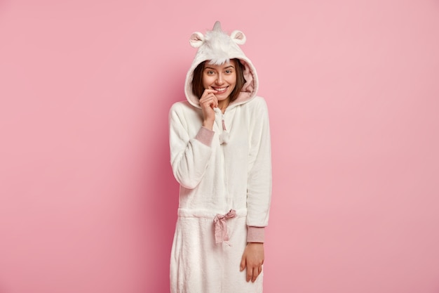 Chica despreocupada con apariencia europea, viste un traje blanco suave kigurumi, mantiene el dedo índice en los labios, se para contra la pared rosada, tiene tiempo libre en casa. Personas, emociones, concepto de estilo de vida