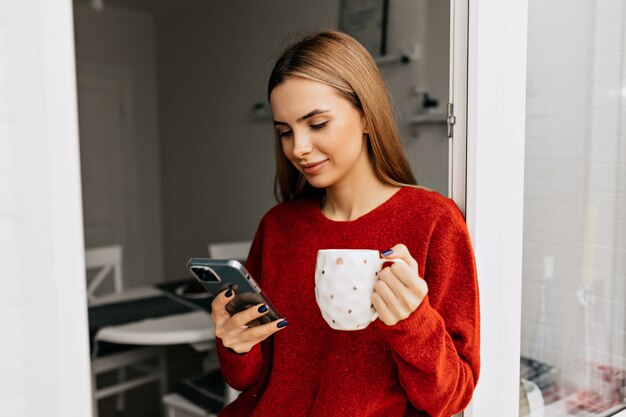 Chica despreocupada bebiendo té en el balcón y usando el teléfono inteligente. Foto de mujer agradable en suéter rojo de punto disfrutando de un café.