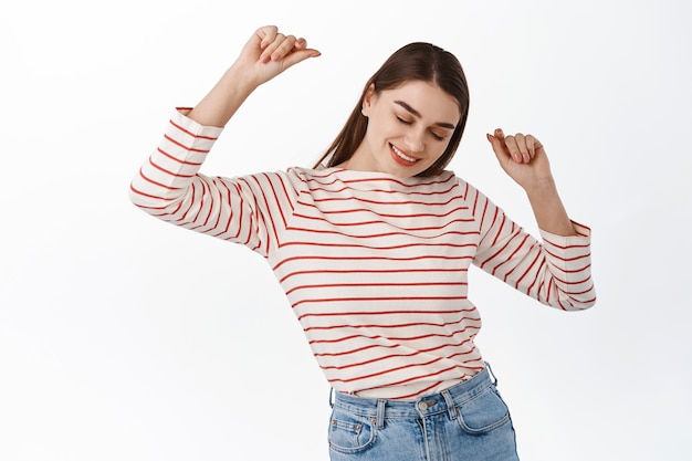 Foto gratuita chica despreocupada disfrutando del tiempo libre, levantando las manos y bailando, sonriendo alegre, divirtiéndose, posando optimista en ropa casual contra el fondo blanco, celebrando