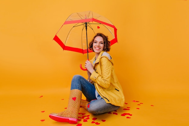 Foto gratuita chica despreocupada en elegante traje de otoño posando con las piernas cruzadas bajo la sombrilla. retrato de interior de dama encantadora con paraguas riendo en pared amarilla.