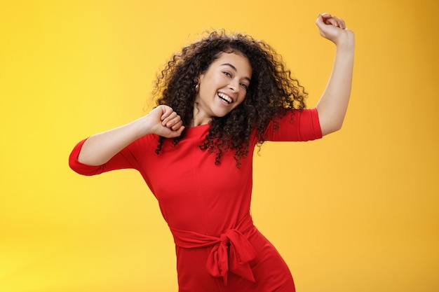 Chica despreocupada que se siente elevada y alegre bailando en vestido rojo levantando las manos felizmente inclinando la cabeza y sonriendo ampliamente a la cámara mientras disfruta de las vacaciones, celebrando las fiestas sobre fondo amarillo.