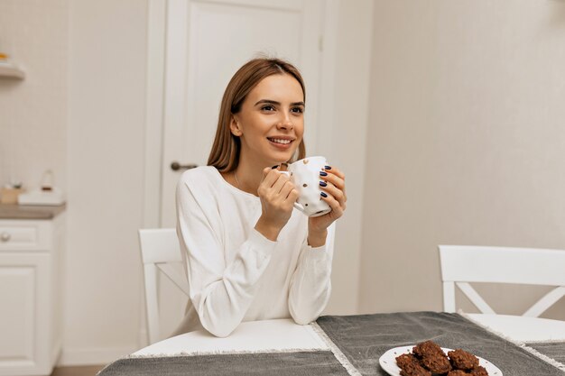 Chica despreocupada tomando café en la cocina. Foto de mujer sonriente agradable en camisa blanca disfrutando de una pausa para el café.