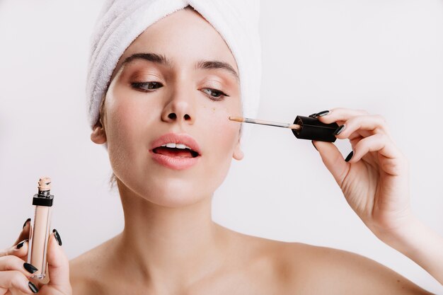 Chica después de la ducha se puso corrector debajo de los ojos. Retrato de mujer hermosa en pared blanca.