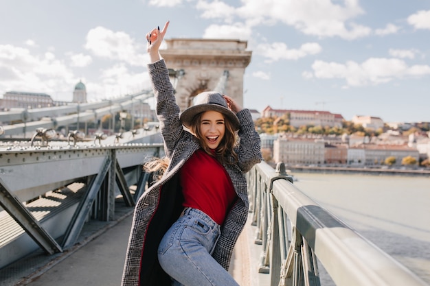 Chica dichosa bien formada en suéter rojo bailando en el puente sobre fondo borroso de la ciudad en la mañana de otoño