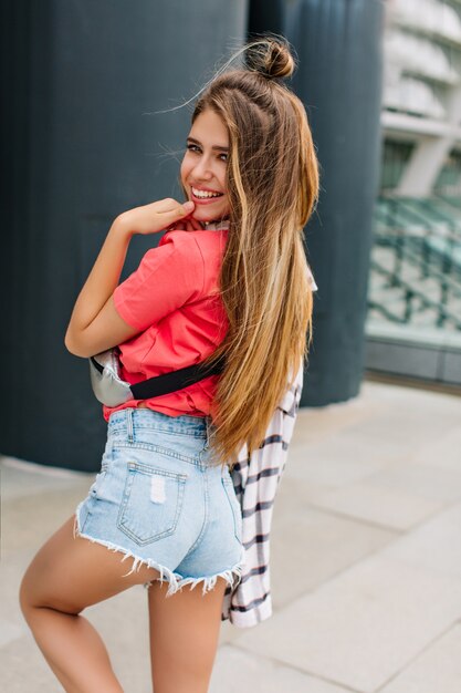 Chica dichosa con peinado de moda en pantalones cortos de mezclilla bailando con hermosa sonrisa y mirando por encima del hombro