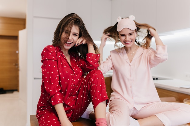 Chica dichosa en ropa de dormir rosa sentada con las piernas dobladas y sonriendo. Retrato de interior de dama morena alegre en ropa roja posando en la cocina.