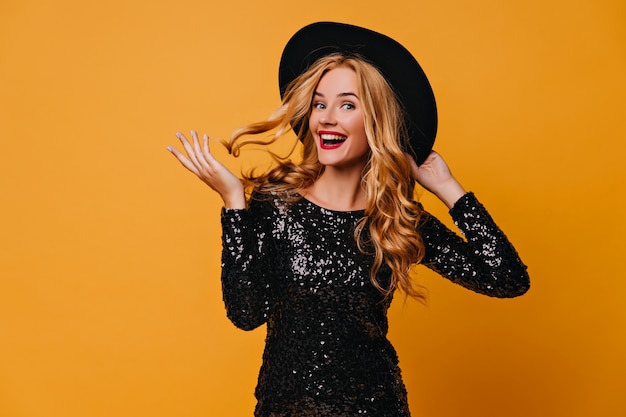 Chica dichosa en vestido negro jugando con su pelo largo. Foto interior de mujer caucásica entusiasta lleva sombrero.