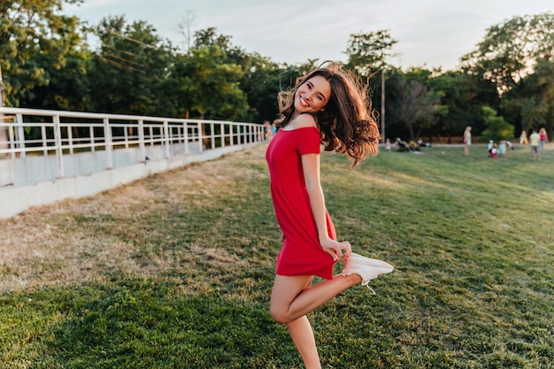 Chica dichosa en vestido rojo divertido bailando en la hierba. Mujer morena refinada riendo durante la sesión de fotos en el parque en un día cálido.