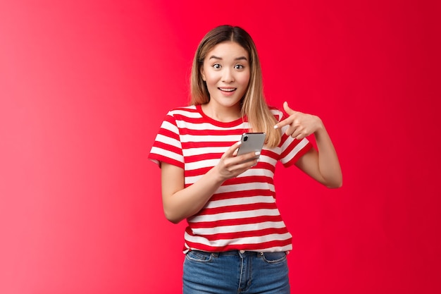 Chica diciéndole acerca de la tienda en línea de promoción impresionante sonriendo impresionado sostenga el teléfono inteligente apuntando con el dedo p ...