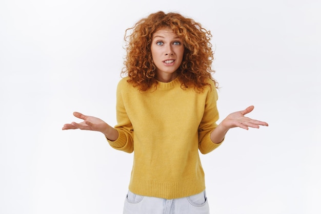 Chica disculpándose encogiéndose de hombros porque no sabe que no puede ayudar Linda mujer pelirroja tonta e incómoda con suéter amarillo extendió los brazos hacia los lados sonriendo torpe y avergonzada admitir su culpa fondo blanco