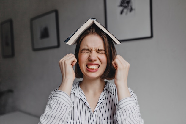 Chica disgustada con camisa a rayas está enojada con los ojos cerrados y el libro abierto en la cabeza contra el fondo de la habitación con imágenes.