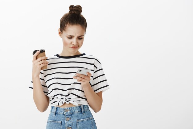 Chica disgustada comprando en línea en la aplicación de teléfono móvil, desplazándose por los perfiles de la aplicación de citas y sonriendo decepcionada mientras bebe café