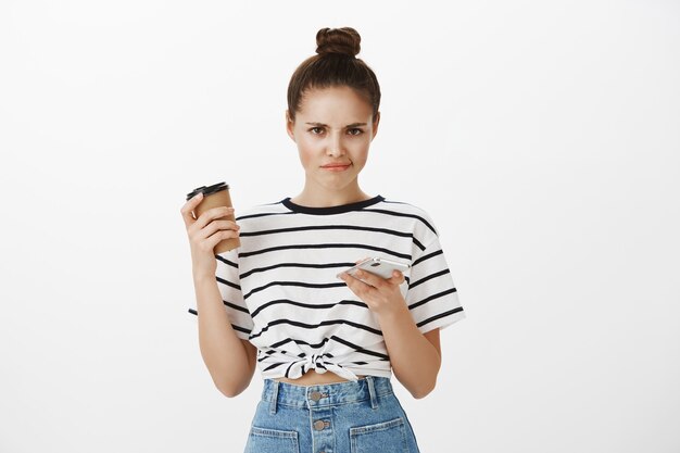 Chica disgustada comprando en línea en la aplicación de teléfono móvil, desplazándose por los perfiles de la aplicación de citas y sonriendo decepcionada mientras bebe café
