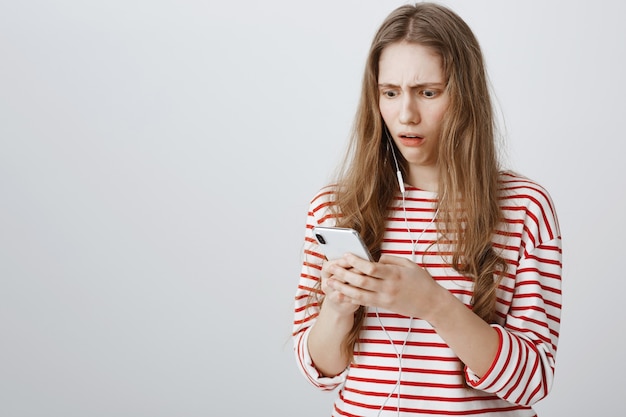 Chica disgustada y sorprendida mira fijamente la pantalla del teléfono móvil preocupada
