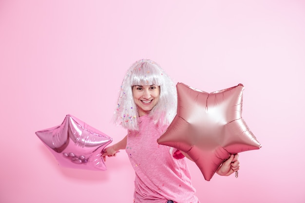 Chica divertida con cabello plateado da una sonrisa y emoción sobre fondo rosa. Joven mujer o niña adolescente con globos y confeti