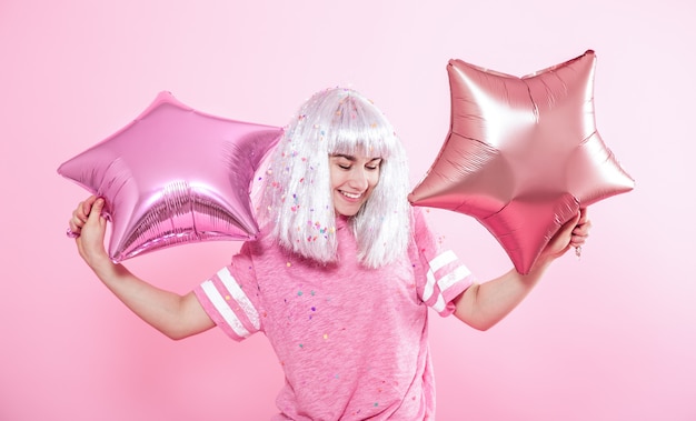 Chica divertida con cabello plateado da una sonrisa y emoción sobre fondo rosa. Joven mujer o niña adolescente con globos y confeti