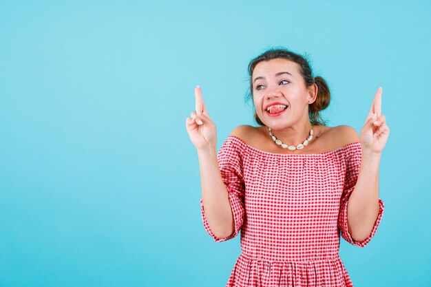 Chica divertida está sacando la lengua y apuntando hacia arriba con los dedos índices sobre fondo azul.