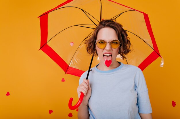 Chica divertida en gafas de sol vintage mostrando la lengua durante la sesión de fotos en la pared amarilla. Foto interior de mujer rizada en camiseta azul de pie bajo una sombrilla.