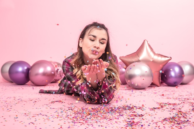 Chica divertida sopla confeti en sus manos. Globos de colores sobre un fondo rosa. El concepto de fiesta y vacaciones.