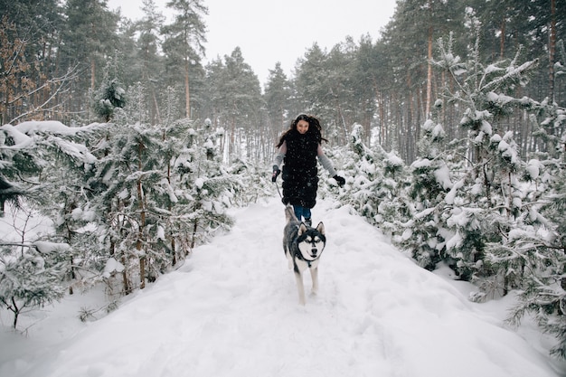 Chica divertirse con su perro Husky en el bosque de pinos de invierno cubierto de nieve