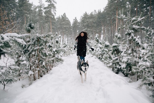 Chica divertirse con su perro Husky en el bosque de pinos de invierno cubierto de nieve