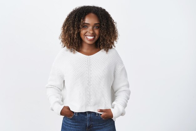 Chica divirtiéndose hablando con una persona interesante Retrato de una joven afroamericana alegre, carismática y sonriente con el pelo rizado en un suéter cogido de la mano en los bolsillos y sonriendo ampliamente a la cámara