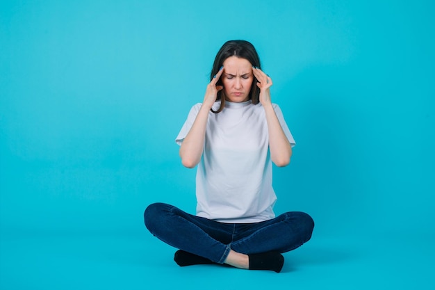 Chica con dolor de cabeza está sosteniendo las manos en las sienes y sentada en el suelo sobre fondo azul.