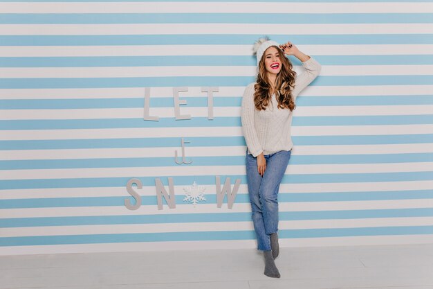 Chica dulce y benevolente con peinado largo y lujoso sonríe felizmente en la pared rayada. Retrato de modelo europeo en sombrero de invierno en pleno crecimiento