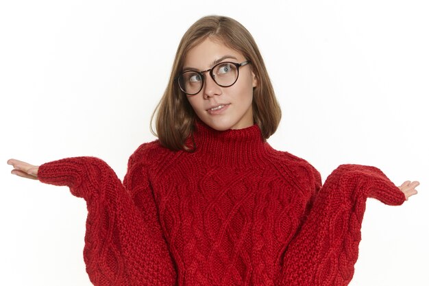 Chica elegante en anteojos y suéter encogiéndose de hombros, perdida, no sabe cómo responder, mirando hacia arriba con expresión facial indiferente despistada. Woamn joven confundido posando