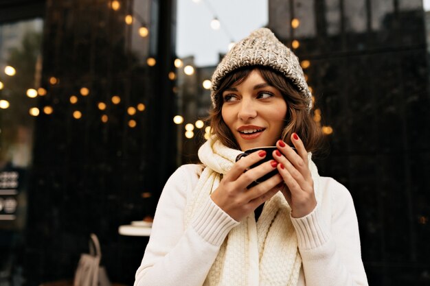 Chica elegante y atractiva en suéter blanco y gorro de punto tomando café afuera en el fondo de la ciudad con luces Foto de alta calidad