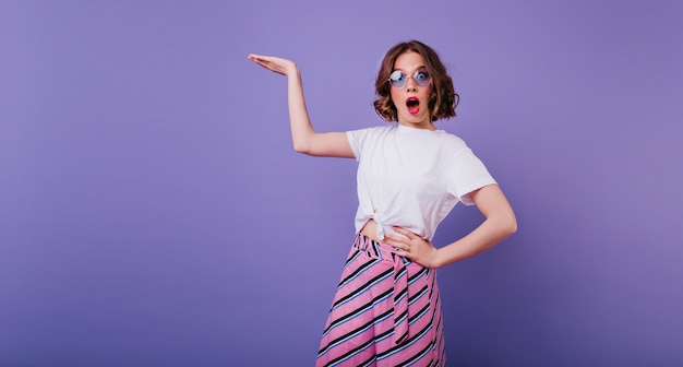 Chica elegante con cabello castaño ondulado que expresa emociones de sorpresa durante la sesión de fotos. Retrato de interior de mujer caucásica sorprendida en vasos de pie en la pared púrpura con la mano hacia arriba